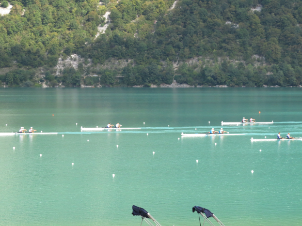 Eine Gruppe von Menschen, die auf einem Gewässer umgeben von Bäumen und einem Hügel rudern, mit Bekleidung-umhangenen Tribünen unten, was auf einen Ruderwettbewerb hinweist.