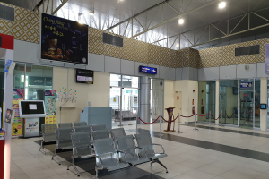 Leerer Flughafen-Wartebereich mit Stühlen auf dem Boden, Absperrpoller mit Seilen, Informationsanzeigen, wandmontierte Bildschirme, Deckenleuchten, Glastüren und andere Einrichtungsgegenstände.