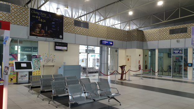 Leerer Flughafen-Wartebereich mit Stühlen auf dem Boden, Absperrpoller mit Seilen, Informationsanzeigen, wandmontierte Bildschirme, Deckenleuchten, Glastüren und andere Einrichtungsgegenstände.