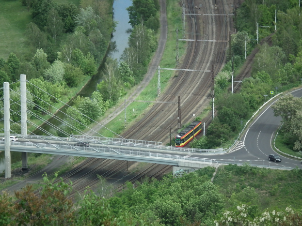 Ein Zug fährt über eine Brücke über einen Fluss, mit Fahrzeugen auf der Straße darunter, Strommasten und -leitungen neben der Brücke und Bäumen und Gras im Hintergrund.