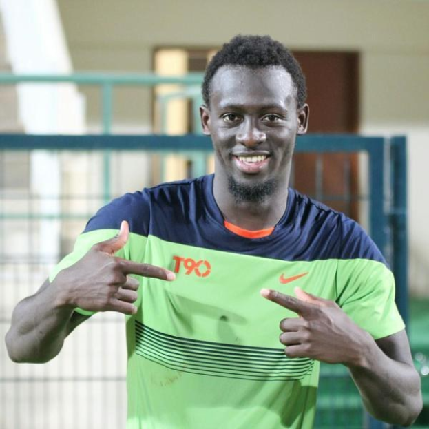 Ein Mann in einem grünen und blauen Hemd, der lachend in die Kamera zeigt, mit einem unscharfen Geländer, einer Wand und einer Tür im Hintergrund.