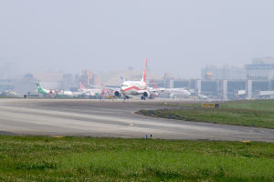 Flughafens Rollbahn mit mehreren geparkten Flugzeugen, grünem Umfeld, Gebäuden im Hintergrund und einem klaren blauen Himmel.