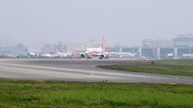 Flughafens Rollbahn mit mehreren geparkten Flugzeugen, grünem Umfeld, Gebäuden im Hintergrund und einem klaren blauen Himmel.