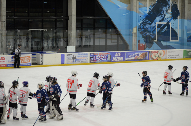 Gruppe junger Menschen, die Eis Hockey auf einer Indoor-Eisfläche spielen, mit Helmen und Stöcken, sowie einer Person auf der linken Seite und Bannern, Säulen und Glaswänden im Hintergrund.