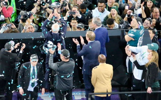 Eine Gruppe von Philadelphia Eagles Spielern feiert mit dem Vince Lombardi Trophy nach dem Gewinn des Super Bowls, einige tragen Mützen und andere werfen Konfetti, während Kameras den Moment festhalten.