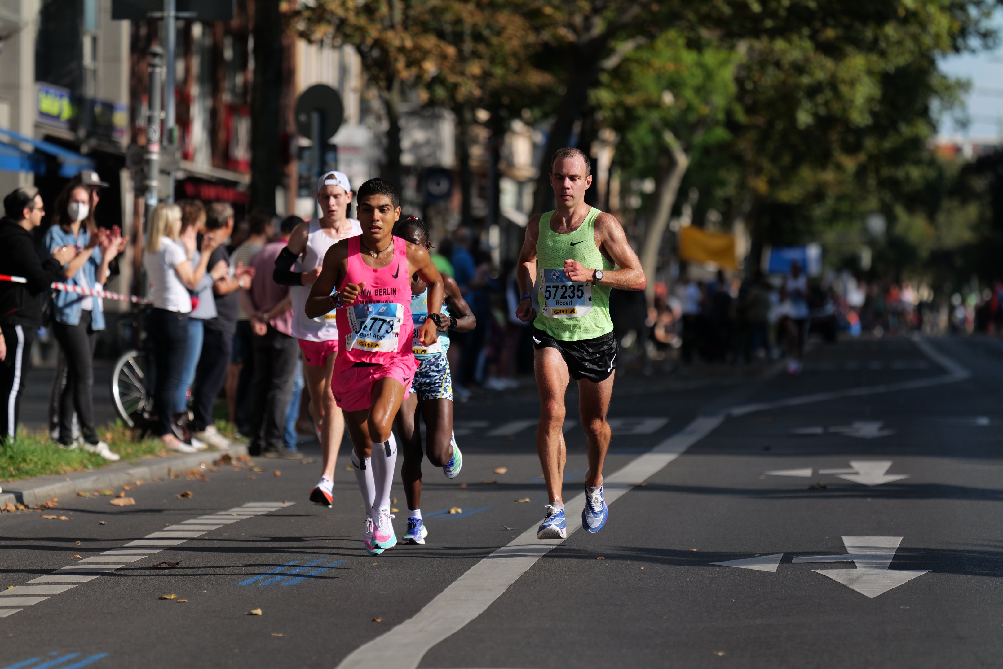 Gruppe von Menschen, die bei einem Marathon auf einer Straße laufen, mit Zuschauern auf der linken Seite, unscharfen Hintergrund mit Bäumen, Gebäuden und einem Fahrrad.