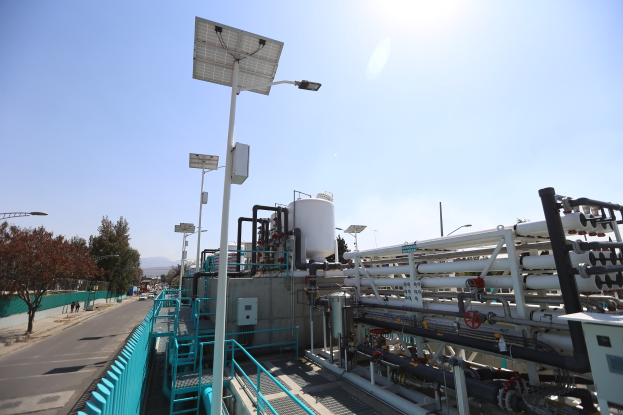 Großes industrielles Gebäude mit umfangreichen Rohrleitungen und Solarpaneelen auf dem Dach, umgeben von Geländern, Polen, Schildern, Bäumen und Fahrzeugen auf der Straße; Teil von Kaliforniens neuer Wasseraufbereitungsanlage.