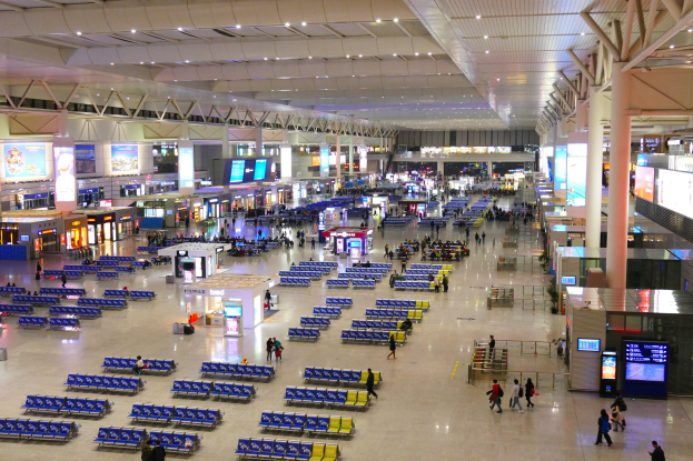 Innenansicht eines Flughafen-Terminals mit Menschen, blauen Stühlen, Geschäften, Informationsanzeigen, Bildschirmen, tragenden Säulen, Beleuchtung und verschiedenen Einrichtungsgegenständen.