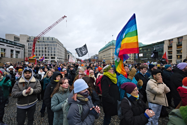 Eine große Gruppe von Menschen mit Fahnen und Schildern vor einem Gebäude während einer LGBTQ+-Rechtsdemo in Berlin, mit einem Kran und Gebäuden im Hintergrund.