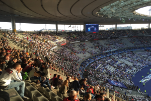 Große Menschenmenge in einem Stadion bei einem Fussballspiel, mit einer Bühne rechts, Fahnen, Stangen, einem Bildschirm und der Allianz Arena in München, Deutschland im Hintergrund.