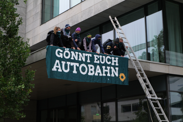 Eine Gruppe von Menschen auf einem Gebäude mit einem Banner, einem Baum links und Glasfenstern im Hintergrund, wahrscheinlich protestierend für die deutsche Regierung.