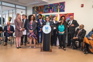 Gruppe von Menschen in einem Raum mit einem Podium, Mikrofon und einer Tafel mit der Aufschrift "Sancho City Councilman Joins City Council", umgeben von Flaggen, Stühlen und Deckenleuchten.