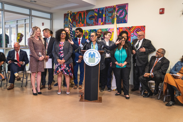 Gruppe von Menschen in einem Raum mit einem Podium, Mikrofon und einer Tafel mit der Aufschrift "Sancho City Councilman Joins City Council", umgeben von Flaggen, Stühlen und Deckenleuchten.