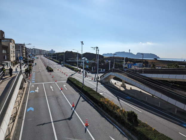 Straße mit Verkehrskegeln, eine Brücke mit Geländern und Laternen, Gebäude mit Fenstern und einige Menschen, vor Bergen und einem bewölkten Himmel, Teil des Takayama-Brückenprojekts.