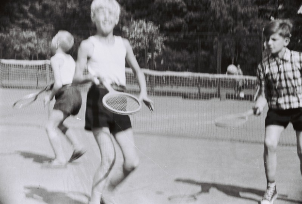 Schwarzes und weißes Foto von drei Kindern auf einem Tennisplatz, die Rackets halten, mit einem Netz, Menschen und Bäumen im Hintergrund.