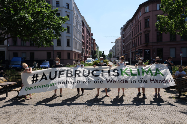 Eine Gruppe von Menschen mit Masken, die ein Banner mit der Aufschrift 'Aufbruchsklima' während einer Klimaprotest in Berlin, Deutschland, halten, umgeben von urbanen Elementen wie Bänken, Bäumen und Fahrzeugen.