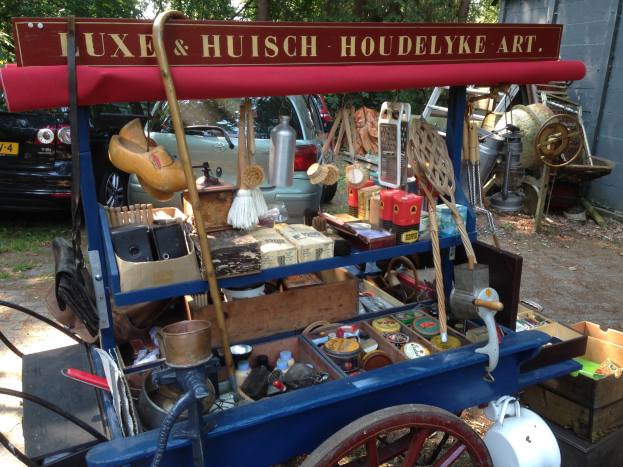 Ein blauer Karren voller Flaschen, Schachteln und anderen Gegenständen steht auf einem Schotterweg auf einem Flohmarkt, mit Fahrzeugen, Bäumen und einer Wand im Hintergrund.