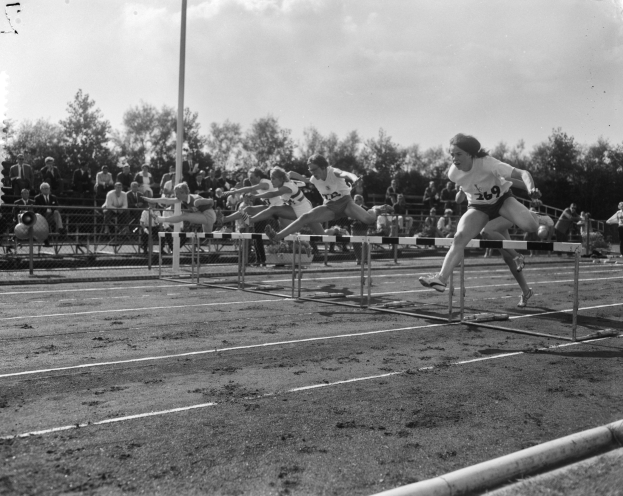 Schwarzes und weißes Foto von Athleten, die über Hürden auf einer Bahn springen, mit einem Pfahl im Vordergrund und Bäumen und Wolken im Hintergrund.