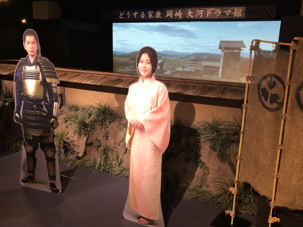 Zwei Menschen in traditioneller japanischer Kleidung - eine Frau in einem pinken Kimono und ein Mann in Samuraikleidung - lächeln gemeinsam in einer Inszenierung mit Pflanzen, einem Schirm und Beleuchtung, die auf eine Filmszene hinweist.