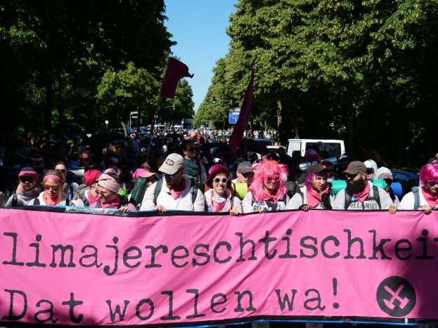 Eine Gruppe von Menschen marschiert die Straße entlang, hölt eine rosa Banner mit 'Women's March in Berlin' darauf, einige tragen Kappen und Schutzbrillen, andere halten Fahnen, mit Bäumen und Fahrzeugen im Hintergrund.
