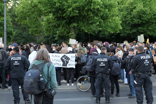 Eine große Gruppe von Menschen bei einer Black Lives Matter Demonstration in Berlin, einige halten Schilder und tragen Mützen und Taschen, mit einem Fahrrad im Vordergrund und Bäumen und einem Pfahl im Hintergrund.