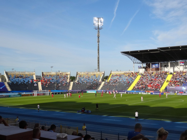 Ein Fußballspiel im Gange in einem großen, beleuchteten Stadion mit Zuschauern auf den Tribünen und Spielern auf dem Feld unter einem sichtbaren Himmel.
