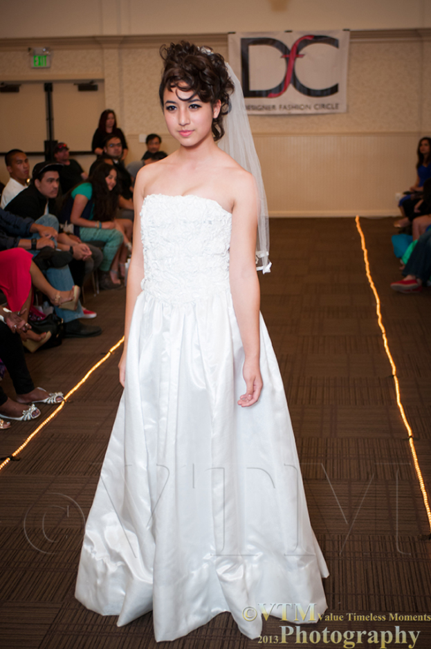 Eine Frau in einem weißen Kleid lächelt und posiert für ein Foto vor einem weißen Banner mit Text darauf, mit Menschen, die auf Stühlen auf beiden Seiten und einem braunen Boden darunter sitzen.