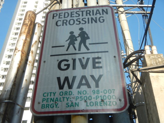 Ein an einem Mast angebrachtes Schild mit der Aufschrift "Fußgängerüberweg Vorrang" umgeben von Strommasten, Drähten, einem fernen Gebäude und Himmel.