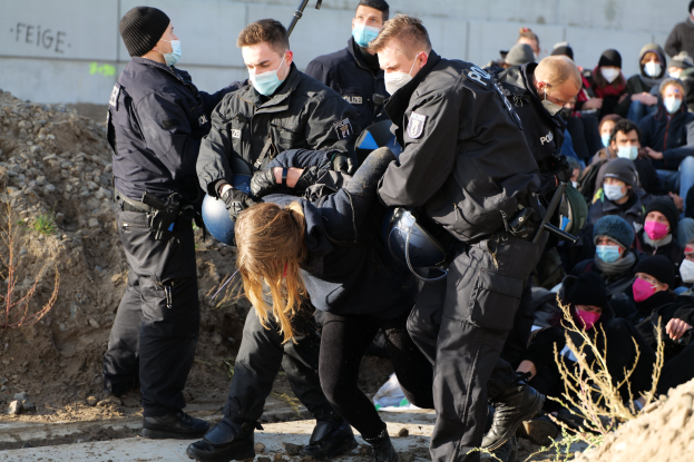 Gruppe maskierter Menschen in Mützen, einige mit Waffen, umgeben eine Frau, die von der Polizei eskortiert wird, in der Nähe von Pflanzen, Steinen und einer Wand mit Protesttext in einer Stadtkulisse.