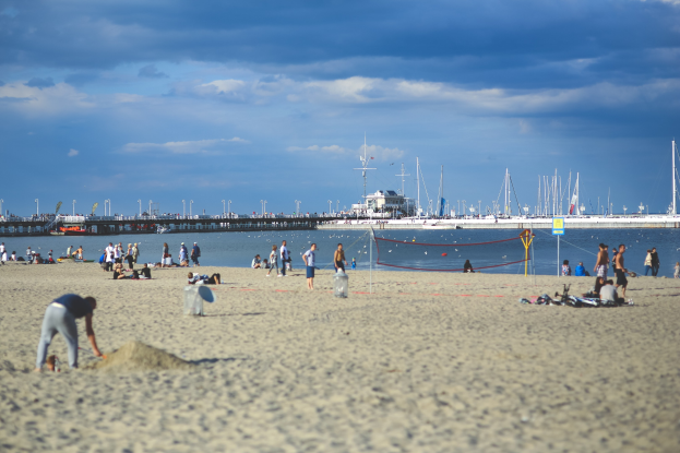 Eine Gruppe von Menschen, die Volleyball auf einem sandigen Strand mit einem Netz spielt, mit Booten, Pfählen und einer Brücke mit Geländern im Hintergrund auf einem großen Gewässer unter einem bewölkten Himmel.