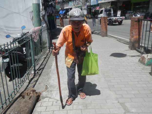Ein älterer Mann geht die Straße entlang, hält einen Stock in einer Hand, eine Tasche in der anderen und einen Stock, trägt eine Mütze, mit einem Metallzaun links und Fahrzeugen, Gebäuden und Schildern im Hintergrund.