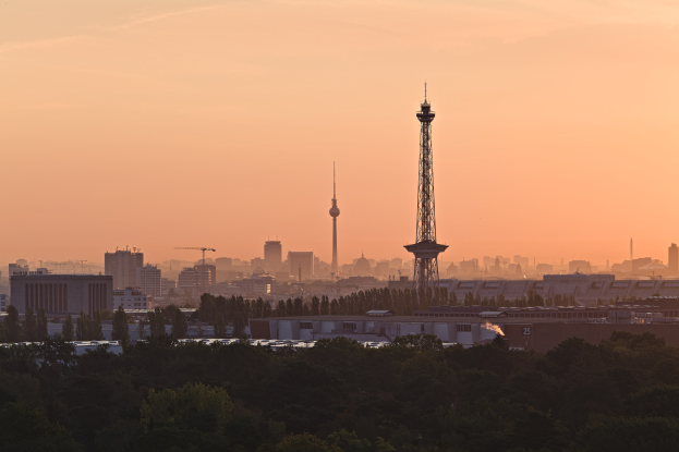 Ein Blick auf die Berliner Skyline bei Sonnenuntergang von einem Hügel aus, mit Gebäuden, Bäumen und dem Fernsehturm, und einem Himmel voller Oranges, Pinks und Purpurs.