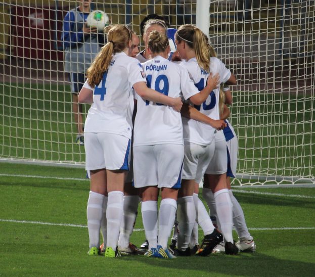 Gruppe von Frauenfußballspielerinnen in Uniformen auf dem Feld mit einem Netz und einer Person, die einen Ball hält, im Hintergrund.