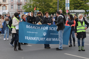 Gruppe von Menschen marschiert die Straße entlang und hält eine 'March for Science Frankfurt am Main'-Fahne mit Bäumen, Pfählen, Schildern, Gebäuden und einem klaren blauen Himmel im Hintergrund.
