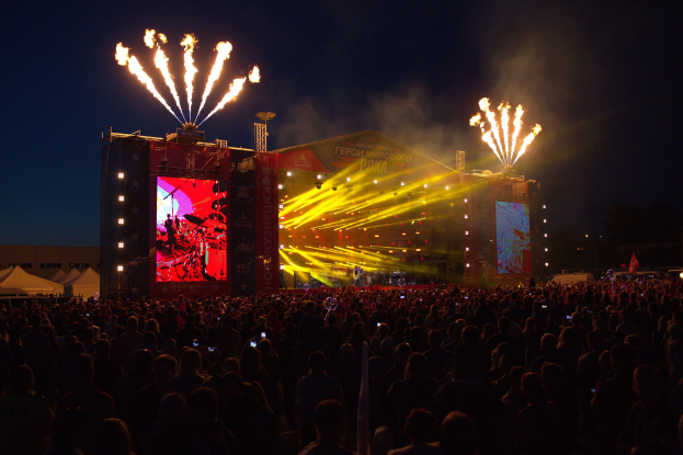 Eine große Menschenmenge auf einem Musikfestival mit beleuchteter Bühne und Performern, umgeben von Bannern, Zelten links und Feuerwerk am dunklen Himmel.