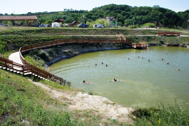 Gruppe von Menschen, die in einem Gewässer mit üppiger grüner Umgebung, einer Brücke mit Geländern und Treppen, Schuppen, Fahrzeugen, Pfählen und einem klaren blauen Himmel im Hintergrund schwimmen.