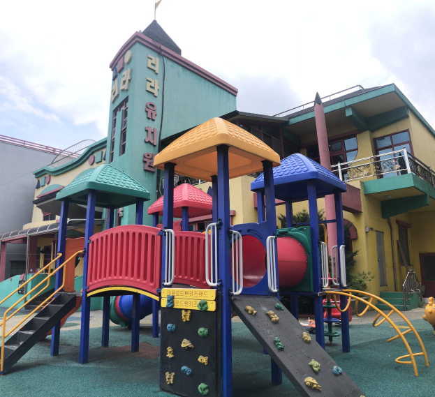 Farbenfrohe Kinderspielplatzgeräte zum Verkauf, mit Rutschen und Kletterstrukturen, vor einem Gebäude mit Fenstern stehend, umgeben von Bäumen unter einem bewölkten Himmel.