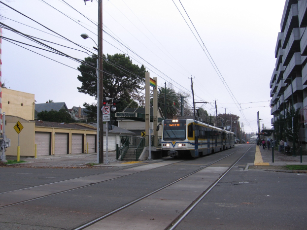 Stadtbahntriebwagen fährt auf einer Straße mit hohen Gebäuden, Strommästen mit Drähten, Passanten auf dem Gehweg, Bäumen und einem klaren blauen Himmel im Hintergrund.