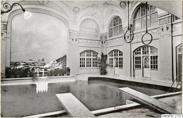 Altes Schwarz-Weiß-Foto eines großen Hallenbads in einem Gebäude mit Glasfenstern und -türen, umgeben von Pflanzen und Deckenleuchten, beschriftet mit "Rensburg, Deutschland - Schwimmbad".