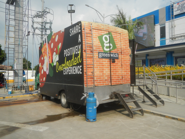 Ein weißer Lkw vor einem Gebäude mit einer Tafel, auf der "Teilen Sie ein positiv überladenes Erlebnis" steht, mit Stufen, einem Zylinder auf dem Boden, Geländern, Strommasten mit Drähten, Laternen, Bäumen und einem bewölkten Himmel.