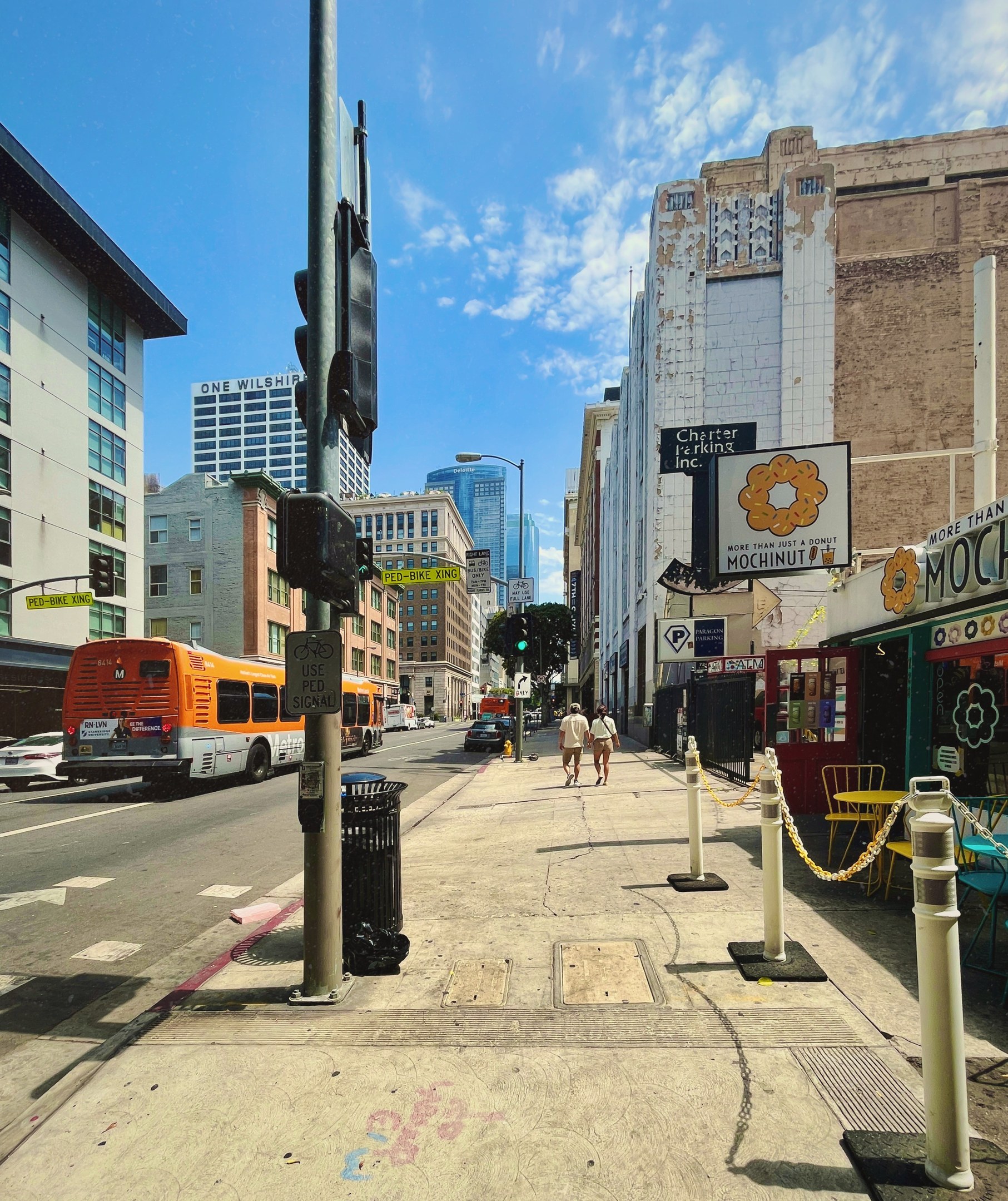 Eine Stadtstraße mit einem an einer Ampel haltenden Bus, Fahrzeugen, Fußgängern, Straßenmöblierung, Gebäuden, Bäumen und einem bewölkten Himmel.