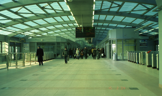 Eine Gruppe von Menschen, die durch einen belebten Bahnhof gehen, mit einem Mann im Vordergrund, umgeben von Geländern, Tafeln mit Text, Lampen und anderen Gegenständen.