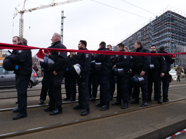 Eine Gruppe von Polizisten in schwarzen Uniformen und Helmen steht in einer Reihe auf einem Bahnsteig, mit einem roten Band davor, während Fahrzeuge, Fußgänger, Gebäude und Strommasten im Hintergrund unter einem klaren blauen Himmel zu sehen sind.