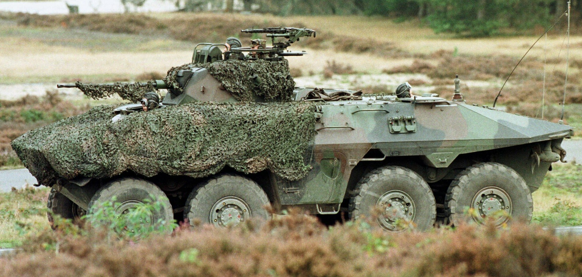 Militärischer Mannschaftstransporter mit montierter großer Kanone, der auf einer Straße neben einem Wald fährt.