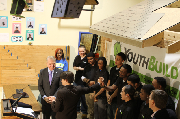 Eine Gruppe von Menschen, die um ein Rednerpult vor einer Menge stehen, wobei einige Hände schütteln, ein Tisch mit Mikrofonen und Papieren links, Plakate an der Wand im Hintergrund und Gegenstände auf dem Dach und der rechten Seite.