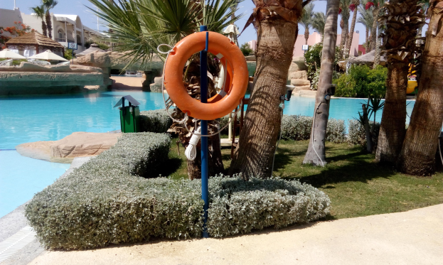 Ein Schwimmbad mit einem Rettungsring in der Mitte, umgeben von üppiger Vegetation, mit Gebäuden, Sonnenschirmen und einem klaren blauen Himmel im Hintergrund.