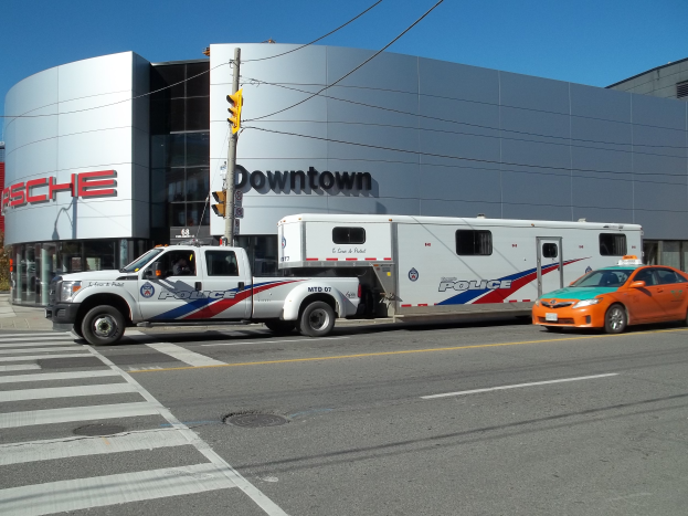 Ein Polizeiwagen vor einem Gebäude mit Text, eine Verkehrsampel mit einem Mast und Drähten im Vordergrund und Bäume mit einem klaren blauen Himmel im Hintergrund.