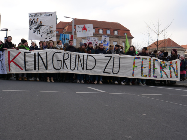 Demonstranten mit einem Transparent mit der Aufschrift "Kein Grund zu Feiern" gegen deutsche Sparmaßnahmen auf einer Straßenkreuzung mit Laternen, Bäumen, Häusern und einem klaren Himmel.