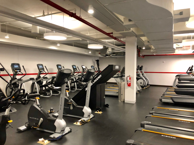 Ein Fitnessraum-Innenraum mit mehreren Laufbändern und Ellipsentrainern in einer Reihe, einer zentralen Säule, einem an der Wand montierten Feuerlöscher und Deckenleuchten in einem Fitnessstudio in New York City.
