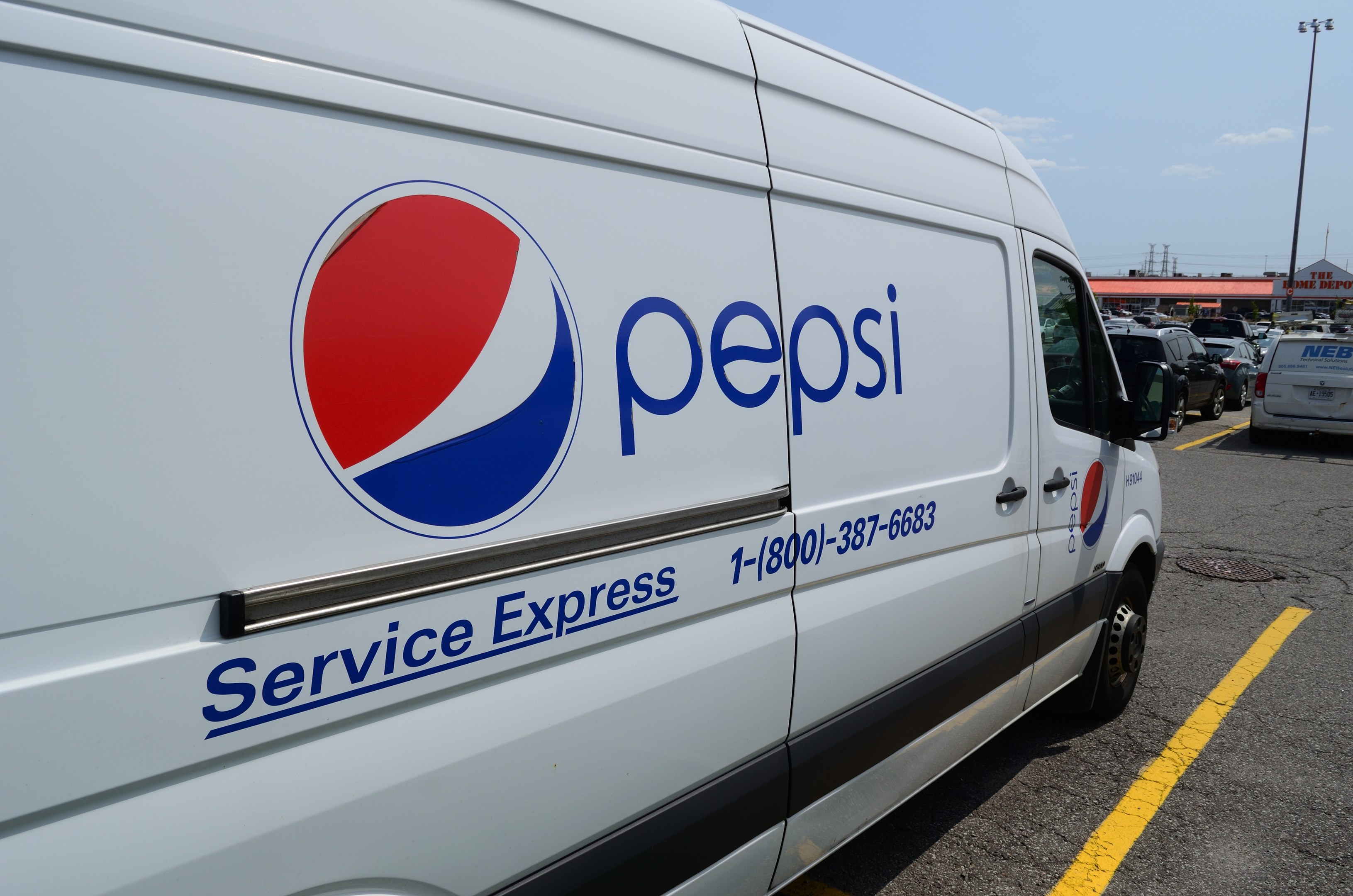 Ein Pepsi Service Express-Lieferwagen auf einem Parkplatz mit anderen Fahrzeugen, Laternenmasten, Gebäuden, Bäumen und einem klaren blauen Himmel im Hintergrund.